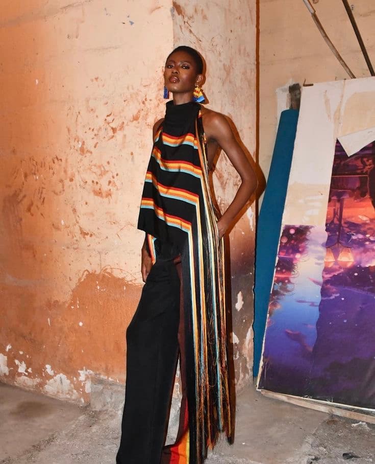Model posing with hand in her pocket, wearing black pants and a sleeveless striped top with that hangs to the ground on one side.